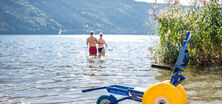 Ein Paar geht im Millstättersee schwimmen, im Vordergrund ein Strandrollstuhl